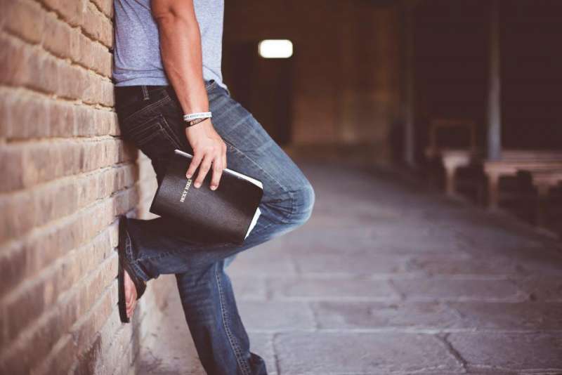 Lower part of a man holding a Bible and leaning against a brick wall